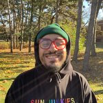 A colour photo shows hip-hop artist standing outside in a field in front of some trees. They are seen smiling wearing a black hoodie with the hood up, red frames, and a green toque. The hoodie says "SUN JUNKIES" in rainbow lettering