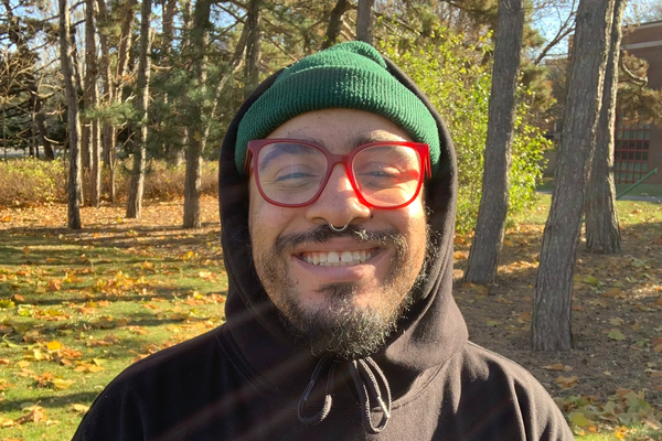 A colour photo shows hip-hop artist standing outside in a field in front of some trees. They are seen smiling wearing a black hoodie with the hood up, red frames, and a green toque. The hoodie says "SUN JUNKIES" in rainbow lettering