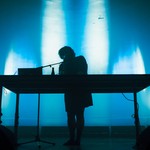 A colour photo shows a silhouette of artist Idlefon standing and performing behind a large table with a laptop on it. The background is a blue lit wall.