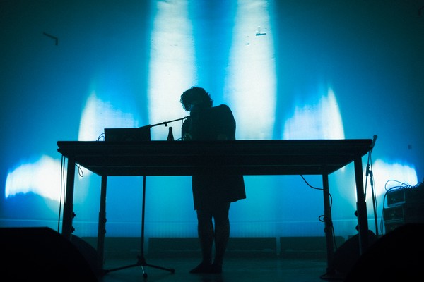 A colour photo shows a silhouette of artist Idlefon standing and performing behind a large table with a laptop on it. The background is a blue lit wall.