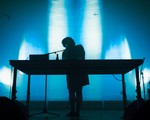 A colour photo shows a silhouette of artist Idlefon standing and performing behind a large table with a laptop on it. The background is a blue lit wall.