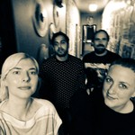 An out of focus sepia tone photo shows four-piece standing in a hallway with posters plastered all over the right side of the wall.
