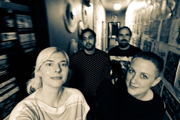 An out of focus sepia tone photo shows four-piece standing in a hallway with posters plastered all over the right side of the wall.