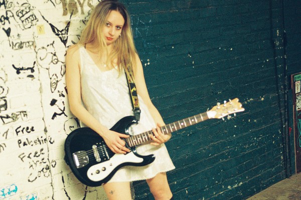 A colour photo of the lead of band Interlay as she stands near a dimly lit skate bowl, leaning against a graffiti-covered wall while holding a black electric guitar. She is wearing a white dress with black knee-high socks and black heels, gazing confidently at the camera.