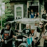 A colour photo shows three members of the rock band JOBSITE are seen playing an outdoor show in front of an old house with people in the background watching.