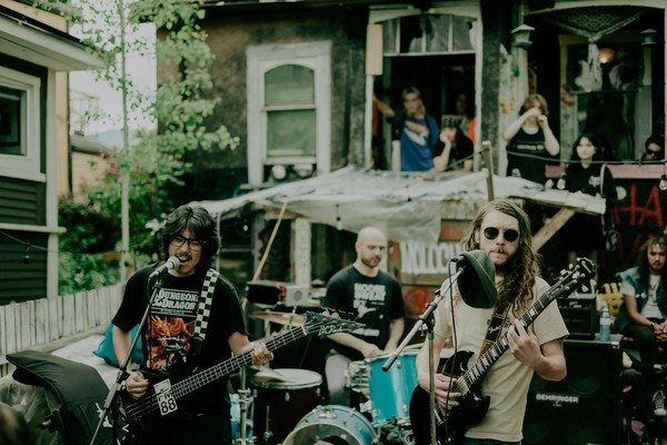 A colour photo shows three members of the rock band JOBSITE are seen playing an outdoor show in front of an old house with people in the background watching.