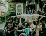 A colour photo shows three members of the rock band JOBSITE are seen playing an outdoor show in front of an old house with people in the background watching.