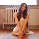 A colour photo shows artist Jolene Marie crouching inside a room wearing a white skirt. A window and a radiator are seen behind them.