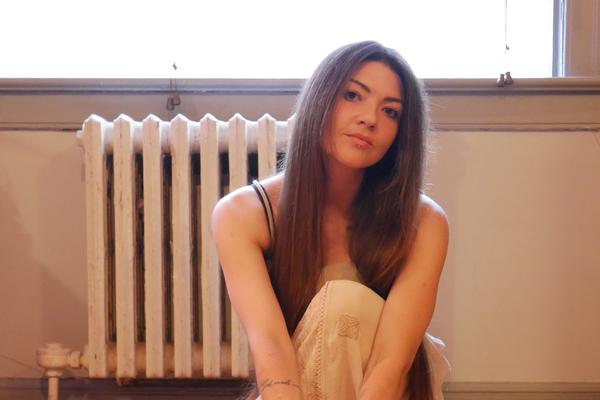 A colour photo shows artist Jolene Marie crouching inside a room wearing a white skirt. A window and a radiator are seen behind them.