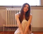 A colour photo shows artist Jolene Marie crouching inside a room wearing a white skirt. A window and a radiator are seen behind them.