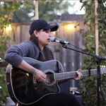 A colour photo shows artist June Hawthorn sitting down and performing live outside, singing into a microphone and playing a black acoustic guitar. They're wearing a black ballcap