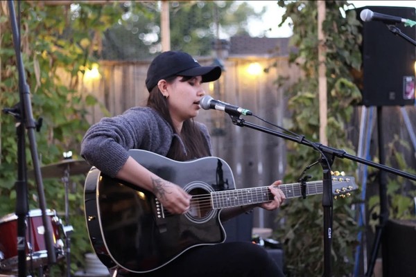 A colour photo shows artist June Hawthorn sitting down and performing live outside, singing into a microphone and playing a black acoustic guitar. They're wearing a black ballcap