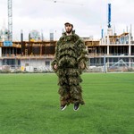 A colour photo shows artist KERUB in a soccer field, jumping in the air wearing a tree-like outfit. An industrial construction site is seen in the background.