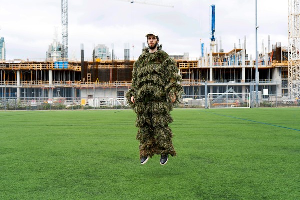 A colour photo shows artist KERUB in a soccer field, jumping in the air wearing a tree-like outfit. An industrial construction site is seen in the background.