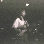 A very grainy black and white photo shows artist KT Laine playing guitar and singing on stage wearing a white top.