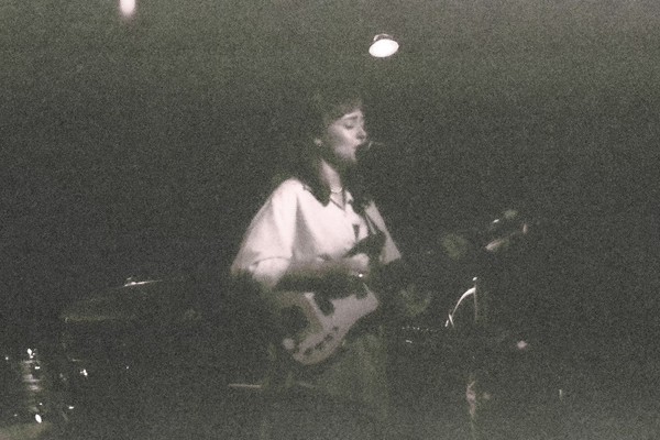 A very grainy black and white photo shows artist KT Laine playing guitar and singing on stage wearing a white top.