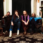 A colour photo shows the four members of Kue Varo & The Only Hopes sitting on a couch in a vintage looking living room with guitar amps on either side and a black velvet curtain hanging in the window behind them.