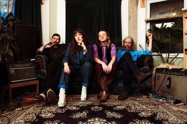 A colour photo shows the four members of Kue Varo & The Only Hopes sitting on a couch in a vintage looking living room with guitar amps on either side and a black velvet curtain hanging in the window behind them.