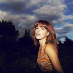 A colour photo shows artist Laraw standing side profile and turning their head toward the camera. An evening sky filled with clouds and a skyline of trees are seen behind them.