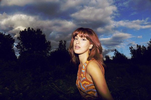 A colour photo shows artist Laraw standing side profile and turning their head toward the camera. An evening sky filled with clouds and a skyline of trees are seen behind them.