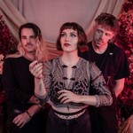 A colour photo shows the three members of Laura Hickli standing in front of a pink backdrop with red leaves and flowers on either side of them.