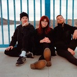 A colour photo shows three-piece group Limited Hangout sitting on the ground of a bridge against a blue railing. You can see a river and trees behind them.