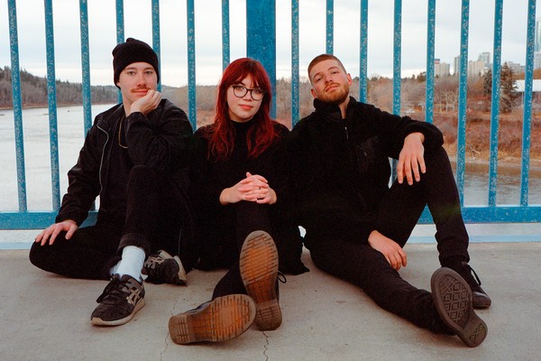 A colour photo shows three-piece group Limited Hangout sitting on the ground of a bridge against a blue railing. You can see a river and trees behind them.