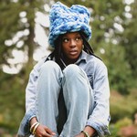 A colour photo shows artist Lowkita sitting with their knees up on a concrete structure with out of focus trees behind them. They're wearing a blue fuzzy hat.