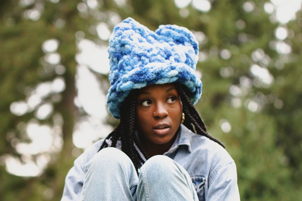 A colour photo shows artist Lowkita sitting with their knees up on a concrete structure with out of focus trees behind them. They're wearing a blue fuzzy hat.