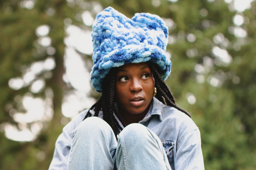 A colour photo shows artist Lowkita sitting with their knees up on a concrete structure with out of focus trees behind them. They're wearing a blue fuzzy hat.
