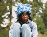 A colour photo shows artist Lowkita sitting with their knees up on a concrete structure with out of focus trees behind them. They're wearing a blue fuzzy hat.