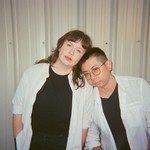 A colour photo shows duo Maneater against a corrugated tin wall. The member on the right rests their head on the shoulder of the other member. Both are wearing open white dress shirts with black t-shirts underneath.