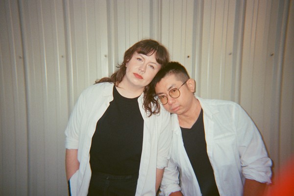 A colour photo shows duo Maneater against a corrugated tin wall. The member on the right rests their head on the shoulder of the other member. Both are wearing open white dress shirts with black t-shirts underneath.