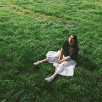 A colour photo taken from above shows artist Marissa Burwell sitting on green grass with a white skirt on looking up at the camera.