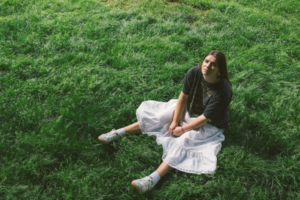A colour photo taken from above shows artist Marissa Burwell sitting on green grass with a white skirt on looking up at the camera.
