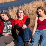 A colour photo of the three members of the band Medusas. The photo shows all three members sitting outdoors in front of a body of water and tall golden reeds. The member of the left is wearing a red jacket and a black t-shirt with white graphics. The member in the middle is wearing a red t-shirt over a black long-sleeve and blue jeans sitting with their arms crossed over their knee. The member on the right wears a red corset-style top and baggy blue jeans with a floral belt, leaning slightly.