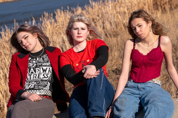 A colour photo of the three members of the band Medusas. The photo shows all three members sitting outdoors in front of a body of water and tall golden reeds. The member of the left is wearing a red jacket and a black t-shirt with white graphics. The member in the middle is wearing a red t-shirt over a black long-sleeve and blue jeans sitting with their arms crossed over their knee. The member on the right wears a red corset-style top and baggy blue jeans with a floral belt, leaning slightly.