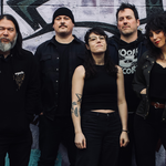 A colour photo of the five members of Midnight Peg. All five members are dressed in all black tops and bottoms, with the middle member wearing a sleeveless top and glasses. They are standing in front of a wall of graffiti.