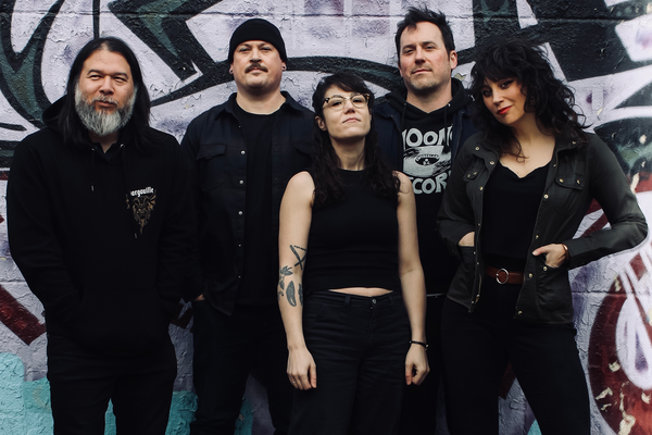 A colour photo of the five members of Midnight Peg. All five members are dressed in all black tops and bottoms, with the middle member wearing a sleeveless top and glasses. They are standing in front of a wall of graffiti.