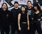 A colour photo of the five members of Midnight Peg. All five members are dressed in all black tops and bottoms, with the middle member wearing a sleeveless top and glasses. They are standing in front of a wall of graffiti.