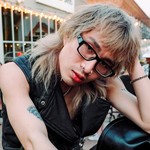 An outdoor portrait of artist MORRISMORRIS shows them sitting with their left hand on their head. They have shaggy blonde hair, black frames and leather vest on.