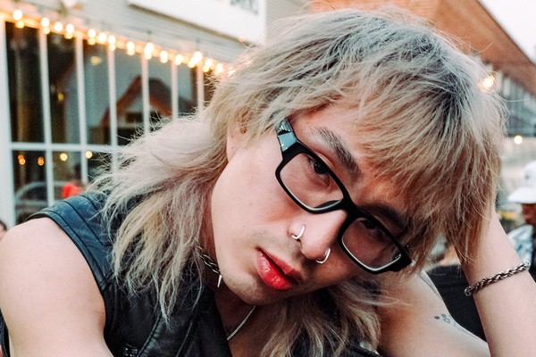 An outdoor portrait of artist MORRISMORRIS shows them sitting with their left hand on their head. They have shaggy blonde hair, black frames and leather vest on.