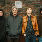 A colour photo shows four-piece band Never Plenty standing in a line in front of a red brick wall. 