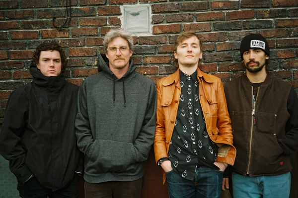 A colour photo shows four-piece band Never Plenty standing in a line in front of a red brick wall. 