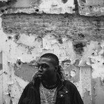 A black and white photo of rapper nk. archaic shows them standing outside in front of a tattered wall wearing a black hooded jacket