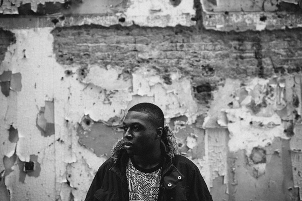 A black and white photo of rapper nk. archaic shows them standing outside in front of a tattered wall wearing a black hooded jacket