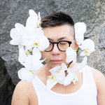 A colour photo of the frontperson of band Non La shows them from the shoulders up wearing a white tank top and black frames. They also have white orchids attached to their face.