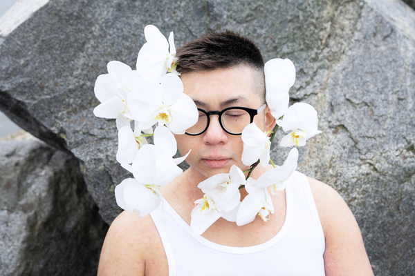 A colour photo of the frontperson of band Non La shows them from the shoulders up wearing a white tank top and black frames. They also have white orchids attached to their face.
