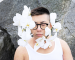 A colour photo of the frontperson of band Non La shows them from the shoulders up wearing a white tank top and black frames. They also have white orchids attached to their face.