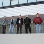 A colour photo of five-piece band standing in a line on a big concrete ledge outside of a building.